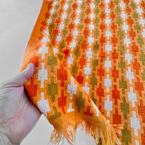May include: A rectangular terry cloth towel featuring a repeating geometric pattern in shades of bright orange, olive green, and off-white. The fabric has a textured surface and is finished with a fringe along the bottom edge of the textile.