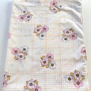 May include: A rectangular fabric pillowcase featuring a white background with a thin, light yellow grid pattern. The surface is decorated with repeating clusters of illustrated flowers in shades of soft pink, white, and muted mustard yellow.