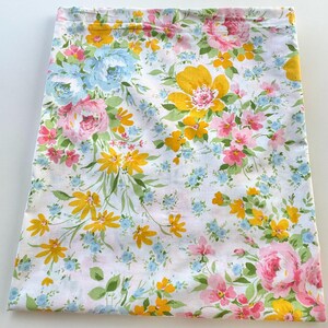 May include: A white fabric with a vibrant floral pattern. The design features large yellow, pink, and blue flowers with green leaves. The fabric is folded, suggesting it could be a tablecloth or a similar textile.