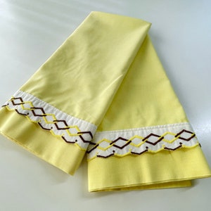 May include: Two pale yellow pillowcases with a decorative embroidered border. The border features a white band with a repeating geometric pattern in brown and yellow. The pillowcases are folded and displayed on a white surface.