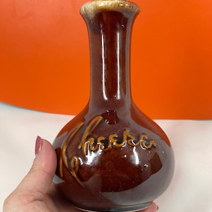 Puede incluir: Un jarrón de cerámica marrón con cuello estrecho y cuerpo redondeado. El jarrón tiene un acabado brillante y un borde marrón claro. La palabra "Cheers" está grabada en la parte delantera en cursiva. El jarrón mide unos 15 cm de alto.