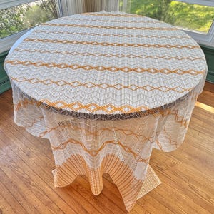 May include: A round table covered with a white and orange patterned lace tablecloth. The tablecloth has a geometric design with horizontal orange stripes and a scalloped edge. The table is set in front of a window.