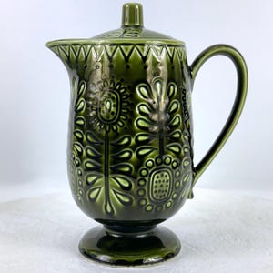 May include: A vintage green ceramic jug with a lid. The jug has a raised floral pattern and a pedestal base. The handle is curved, and the spout is designed for pouring. The design is in a mid-century modern style.
