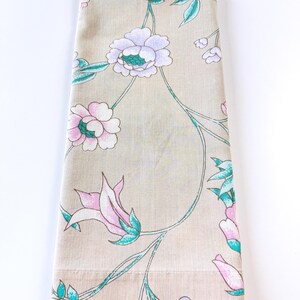 May include: A folded rectangular fabric piece in a light beige tone featuring a repeating floral pattern. The design includes stylized flowers in shades of soft pink, lavender, and white, accented by thin, vibrant teal green stems and leaves.