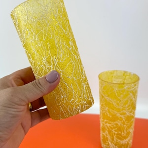 Puede incluir: Dos vasos amarillos con una superficie texturizada y detalles blancos en forma de telaraña. Un vaso es sostenido por una mano, mientras que el otro está sobre una superficie naranja. Los vasos tienen forma cilíndrica.