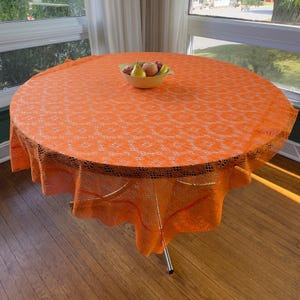 May include: A round, bright orange lace tablecloth with a repeating floral pattern draped over a circular table. Centered on the tablecloth is a light yellow bowl containing a pear, an apple, and a few red grapes, creating a vibrant display.
