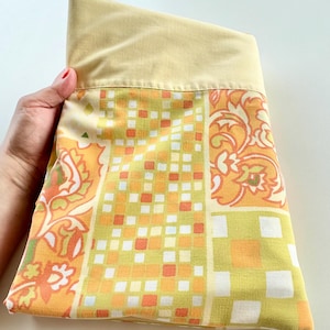 May include: A folded, rectangular pillowcase with a yellow top and a patterned front. The front features a mix of orange, white, and green geometric and floral designs. The pillowcase is held by a hand.