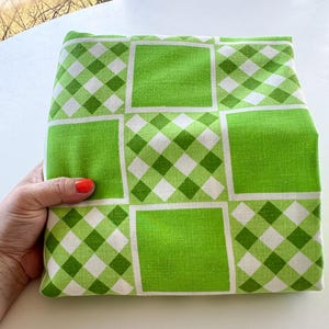 May include: A folded fabric with a green and white checkered pattern. The fabric features large green squares and smaller checkered squares. The fabric is held in a person's hand.