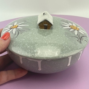 May include: A ceramic trinket box with a light green speckled finish. The lid features a small house with a snow-covered roof and two painted white daisies with yellow centers. White paint drips down the sides of the box.