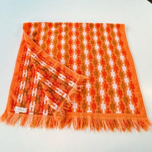 May include: A rectangular orange towel featuring a repeating geometric pattern in white and darker orange. The towel has a fringed edge along the bottom and a small white fabric label in the corner with the text Cannon printed in black.