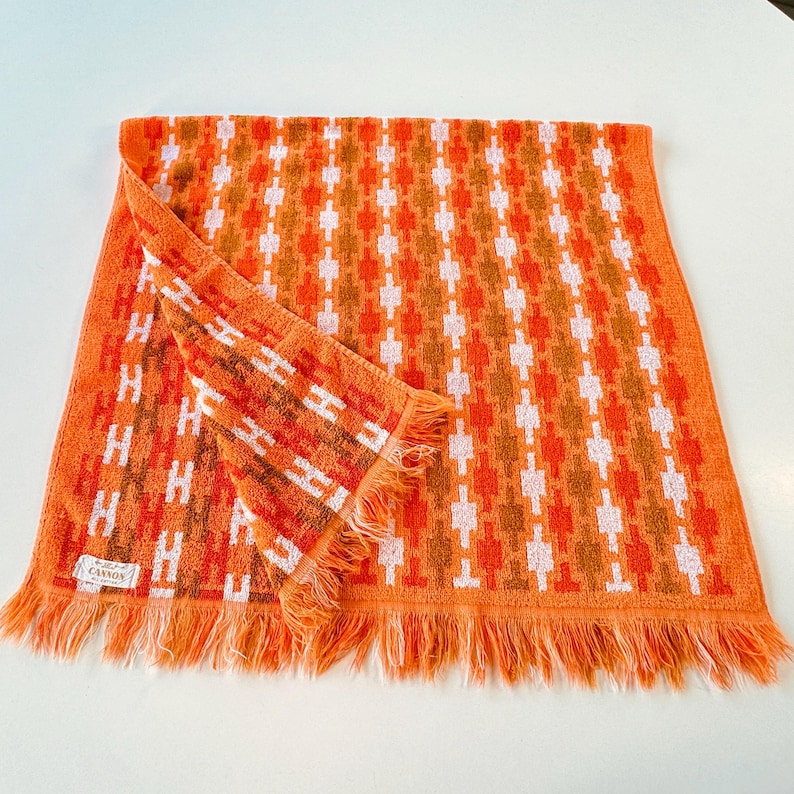 May include: A rectangular orange towel featuring a repeating geometric pattern in white and darker orange. The towel has a fringed edge along the bottom and a small white fabric label in the corner with the text Cannon printed in black.