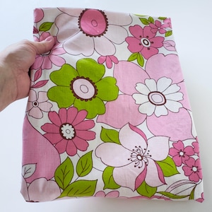 May include: A folded piece of fabric featuring a large-scale floral pattern. The design consists of stylised flowers in shades of light pink, dark pink, and white, interspersed with bright lime green leaves against a white background.