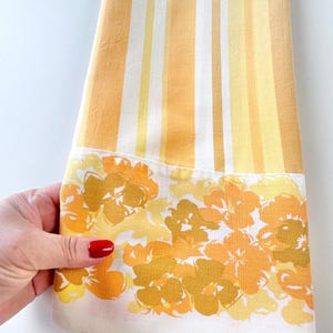 May include: A folded vintage kitchen towel with vertical stripes in shades of yellow and white. The bottom edge features a floral pattern in yellow and orange hues against a white background. The towel is held by a hand with red nail polish.