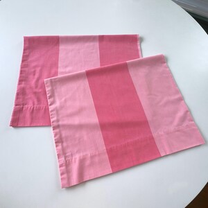 May include: Two rectangular pillowcases featuring a vertical striped pattern in alternating shades of light pink and medium pink. The fabric has a smooth, matte finish with clean, straight stitched hems along the edges of each pillowcase.