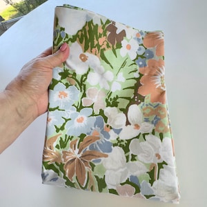 Könnte beinhalten: Ein gefaltetes Stück Stoff mit einem Blumenmuster aus großen weißen, hellblauen und pfirsichfarbenen Blüten vor einem Hintergrund aus Blättern und Blattwerk in verschiedenen Grüntönen. Das Textil zeigt einen dichten, sich wiederholenden botanischen Druck.