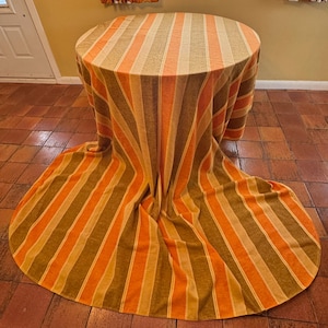 Peut inclure: Une table ronde recouverte d'une nappe à rayures. La nappe présente des rayures verticales dans les tons orange, beige et marron. Le tissu tombe jusqu'au sol, créant un effet fluide. La table est posée sur un sol carrelé.