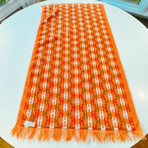 May include: A rectangular orange towel featuring a repeating vertical pattern of white and brown letter H shapes. The towel has a textured terry cloth surface and is finished with a fringed edge on the bottom. A small white label reads Cannon.