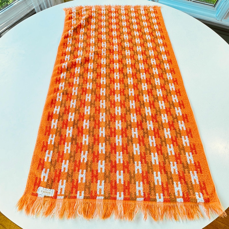 May include: A rectangular orange towel featuring a repeating vertical pattern of white and brown letter H shapes. The towel has a textured terry cloth surface and is finished with a fringed edge on the bottom. A small white label reads Cannon.