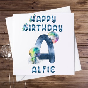 May include: A white birthday card with the text "HAPPY BIRTHDAY ALFIE" in blue. The letter "A" is filled with a starry night design, and a colourful alien and planet are featured. The card is on a wooden surface.