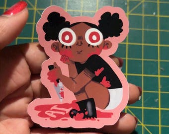 May include: A pink sticker featuring a cartoon illustration of a child with black hair and large red eyes, holding a bloody knife. The sticker has the word "AT" in red blood. The child is wearing a black shirt and white diaper.