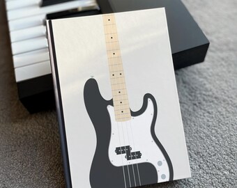 May include: A notebook with a black and white bass guitar illustration. The notebook is resting on a gray surface next to a black keyboard. The bass guitar design features a black body, white pickguard, and a light-colored neck.