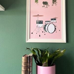 May include: A framed print of a drum set with hanging plants on a pink background. The drum set is black and white with green accents. The frame is white and the background is a green wall.