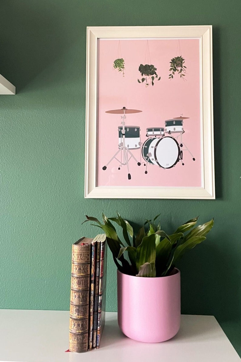 May include: A framed print of a drum set with hanging plants on a pink background. The drum set is black and white with green accents. The frame is white and the background is a green wall.