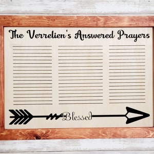 May include: Wooden framed sign with the text "The Verrelien's Answered Prayers". The sign has a light wood background with horizontal lines for writing and a black arrow with the word "Blessed". The frame is a dark brown colour.