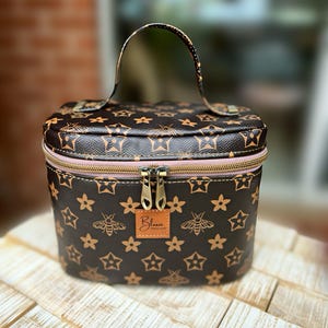 May include: A brown cosmetic bag featuring a pattern of stars, flowers, and bees. It has a pink zip, a handle, and a small tag with the word "Bloom". The bag is placed on a wooden surface.