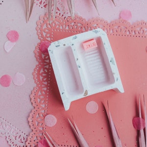 May include: A white ceramic tray with two compartments, one with a pink soap bar that says "LOVE" on it. The tray is on a pink and white doily with pink confetti.