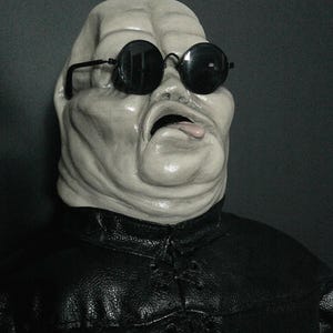 May include: A pale, textured, humanoid mask with a prominent forehead, deep-set features, and an open mouth with a visible tongue. The mask wears round, black-rimmed sunglasses and a black, textured, collared jacket with lace-up detailing.