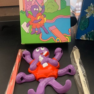 May include: Purple octopus-shaped toothbrush holder with a cartoon octopus holding a toothbrush and toothpaste. The holder has eight tentacles with suction cups. The octopus is wearing a red shirt. The holder is packaged with two toothbrushes.