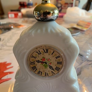 May include: White porcelain clock shaped bottle with a gold top. The clock face has Roman numerals and a floral design with red roses.