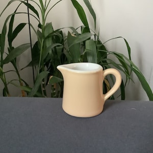 May include: A small, peach-colored ceramic creamer with a white interior and a handle. The creamer is sitting on a gray surface.