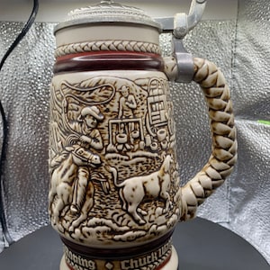 May include: A ceramic beer stein with a brown and white design depicting a cowboy roping a bull. The stein has a metal lid and a braided handle. The text "Chuck Wagon Roping" is printed on the bottom of the stein.