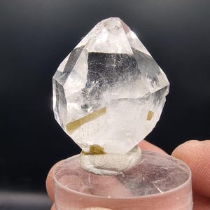 May include: A clear quartz crystal with a geometric shape, mounted on a clear, round base. The crystal has internal inclusions and a polished appearance. The background is dark.