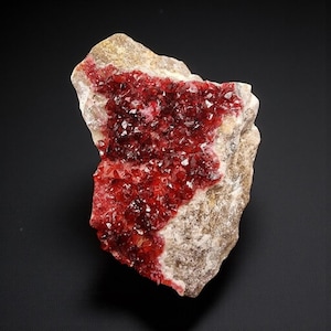 Puede incluir: Espécimen mineral natural. Una matriz de roca beige está cubierta con un grupo de cristales de color rojo intenso. Los cristales están muy juntos, creando un contraste llamativo con el fondo neutral.