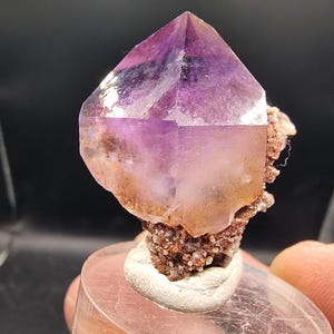Può includere: Cristallo di ametista naturale con punta viola scuro e base traslucida più chiara. Il cristallo è montato su un gruppo di piccole formazioni minerali marrone-rossastre, su una base trasparente di forma ovale.