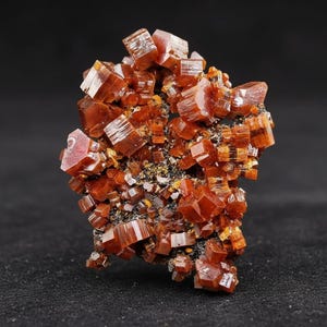 Puede incluir: Primer plano de un espécimen mineral de vanadinita. Los cristales, de un rico tono rojo-anaranjado, están agrupados. Los cristales individuales son prismas hexagonales. El fondo oscuro resalta el color y la textura vibrantes.
