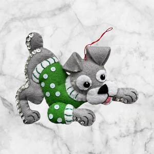 May include: A gray felt dog ornament with a green sweater adorned with white polka dots and white trim. The dog has a black nose, white muzzle, and large, round eyes. Clear sequins are placed along the dog's back and paws. A red string is attached for hanging.