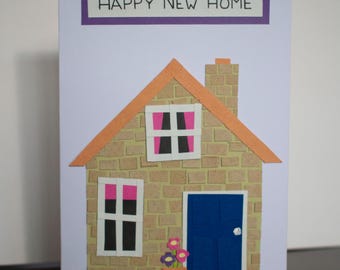 May include: A handmade card with a purple banner that says "Happy New Home". The card features a paper house with a brown brick exterior, a blue door, and a flower pot with pink and purple flowers.