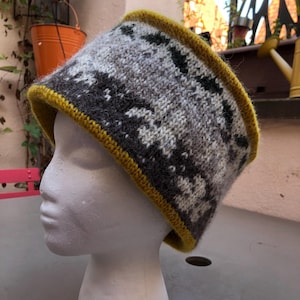 Puede incluir: Una diadema de punto con un patrón geométrico en tonos grises, blancos y negros, acentuada con un ribete amarillo mostaza. La diadema se exhibe en una cabeza de maniquí blanca, mostrando su diseño y textura.