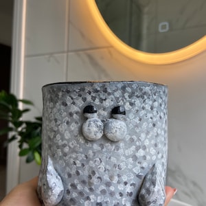 May include: A grey ceramic planter shaped like a seal with white polka dots and black eyes. The planter is being held in a hand.