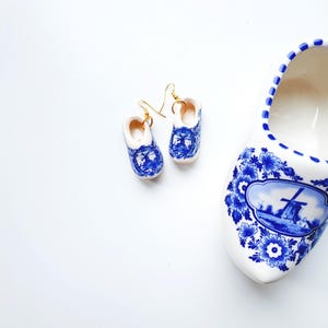 May include: A pair of miniature ceramic clog earrings with gold-toned hooks and a larger ceramic clog, all featuring a white base with blue floral patterns and a detailed windmill illustration on the front of each piece.