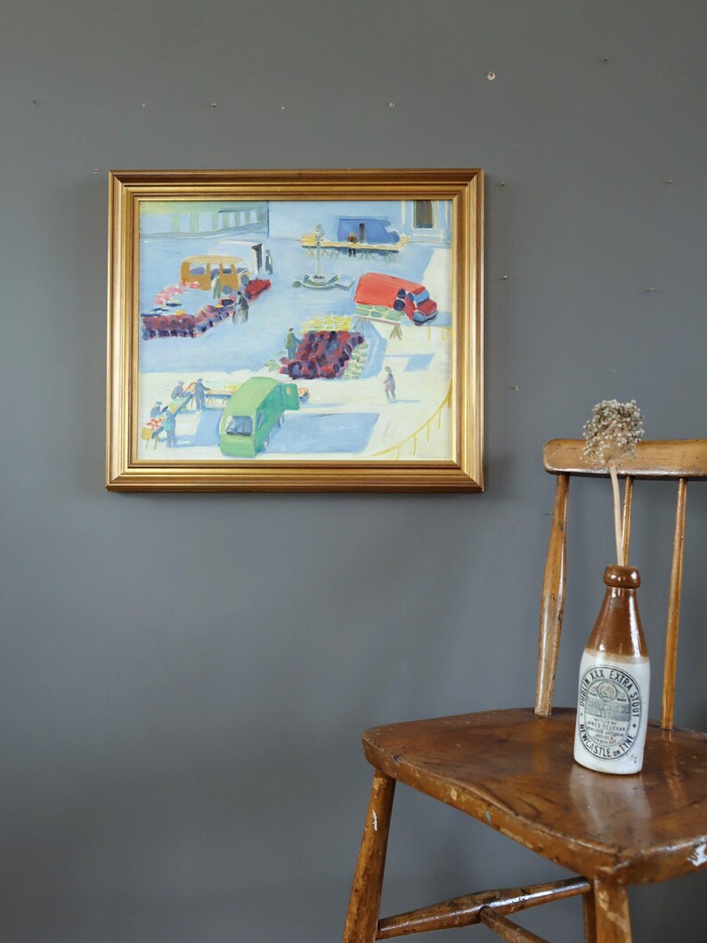 Pode incluir: Uma pintura a &oacute;leo emoldurada mostra uma cena de mercado com ve&iacute;culos coloridos. Uma garrafa vintage castanha e branca com texto est&aacute; numa cadeira de madeira. O fundo &eacute; uma parede cinzenta escura.
