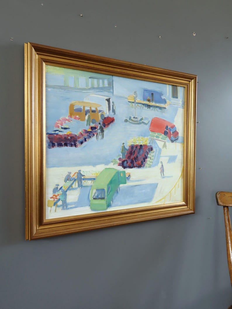 Pode incluir: Uma pintura a &oacute;leo emoldurada em ouro retrata uma movimentada cena de mercado ao ar livre. A obra apresenta ve&iacute;culos coloridos, incluindo um &ocirc;nibus amarelo, um caminh&atilde;o vermelho e uma van verde, rodeados por figuras e bancas de mercado. A pintura est&aacute; montada em uma parede cinza.
