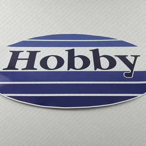 Op de afbeelding: Ovale sticker met blauwe en witte strepen en het woord "Hobby" in zwarte letters.