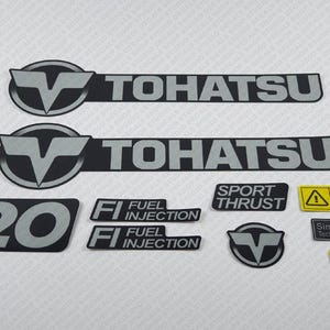 May include: A set of black and silver decals for a Tohatsu outboard motor. The decals include the Tohatsu logo, the number 20, and the text "Fuel Injection" and "Sport Thrust". There is also a yellow decal with a warning symbol and the text "Simpliq Technology".