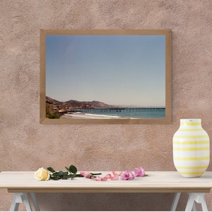 May include: A framed photograph of a wooden pier extending into the ocean with a mountain range in the background. The sky is blue and the water is a light blue. The photo is hanging on a light brown wall.