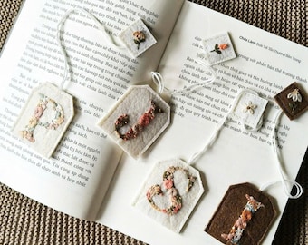 May include: Handmade felt bookmarks with embroidered floral designs and letters. The bookmarks are in various shapes and colors, including white, brown, and beige. They are placed on the pages of an open book, with the text visible.